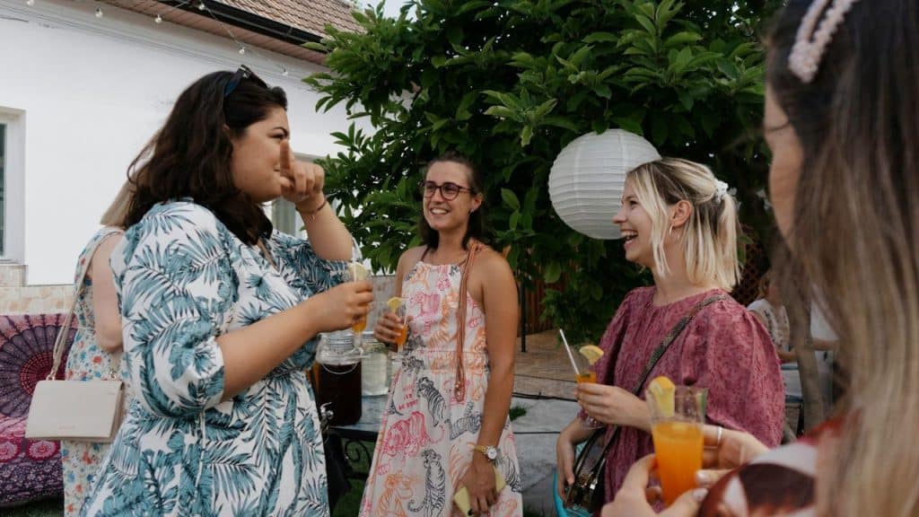 Four women stand outside holding drinks and laughing together near a large green tree.
