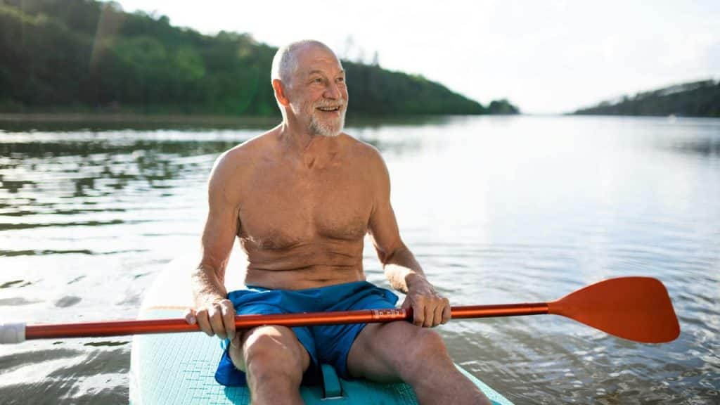 A smiling older man sits on a paddleboard on a lake holding an orange paddle.