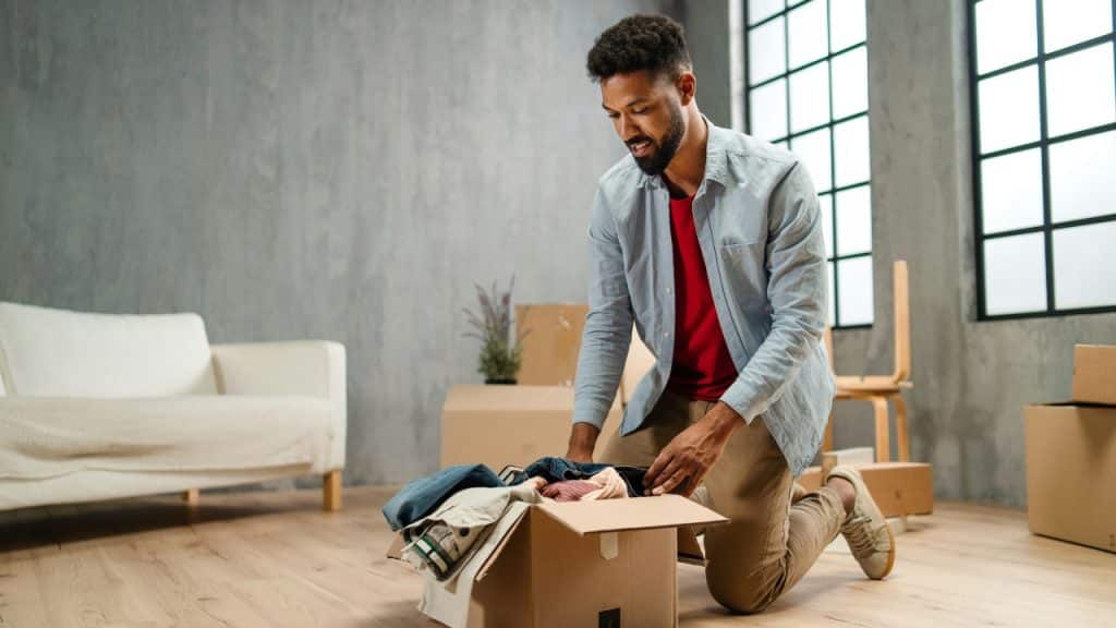 A man kneels on a wooden floor and places folded clothes into a cardboard box.