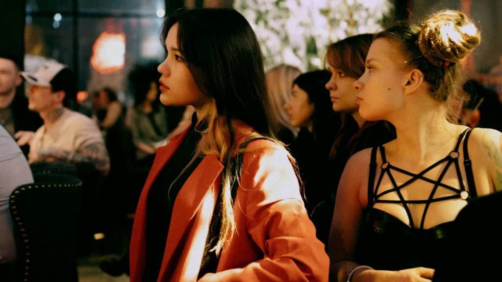 Several people stand in a dimly lit room, with a woman in a red blazer centered.