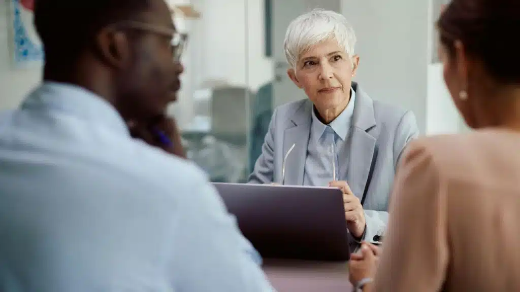 A woman with short grey hair looks at two people sitting across from her at a desk.