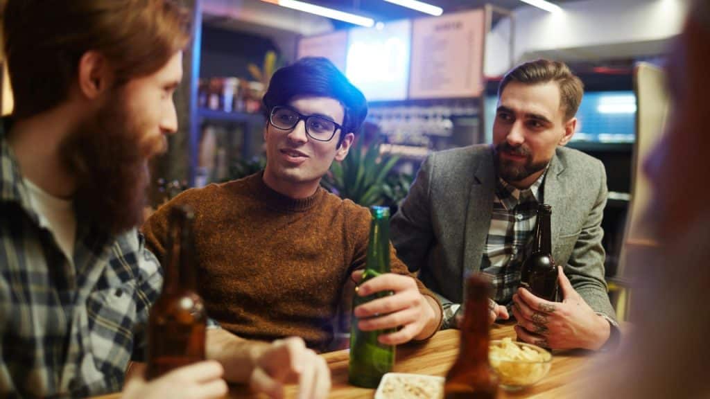 Three men sit at a wooden bar with beer bottles, engaged in a conversation together.