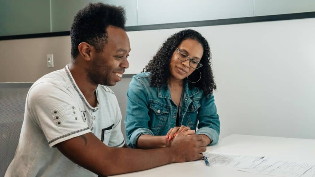 A man and woman sit at a white table, holding hands over a printed document.