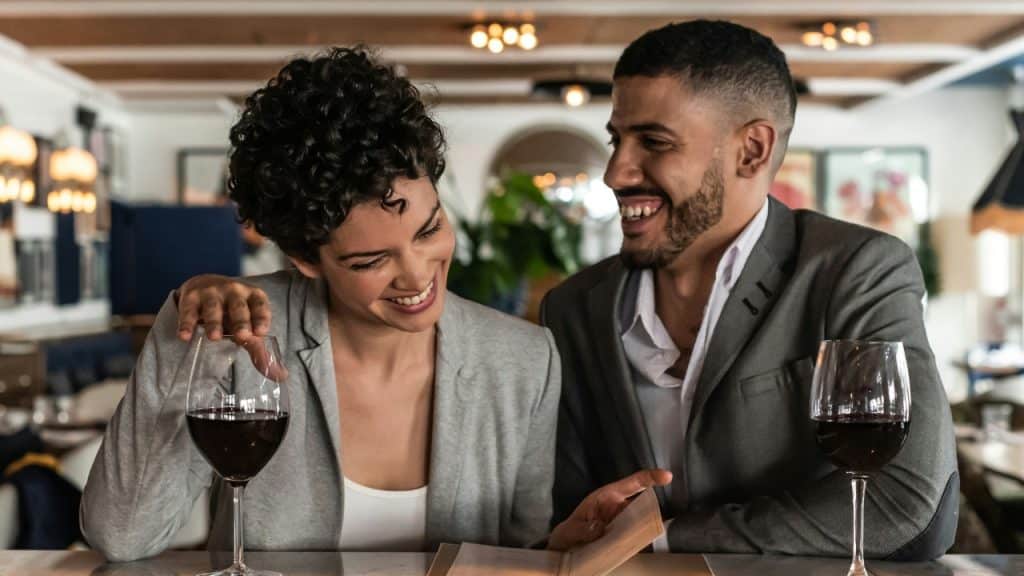 A man and woman laugh while looking at a menu between two glasses of wine.
