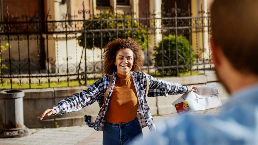 A smiling woman opens her arms while holding a map on a sunny city street.