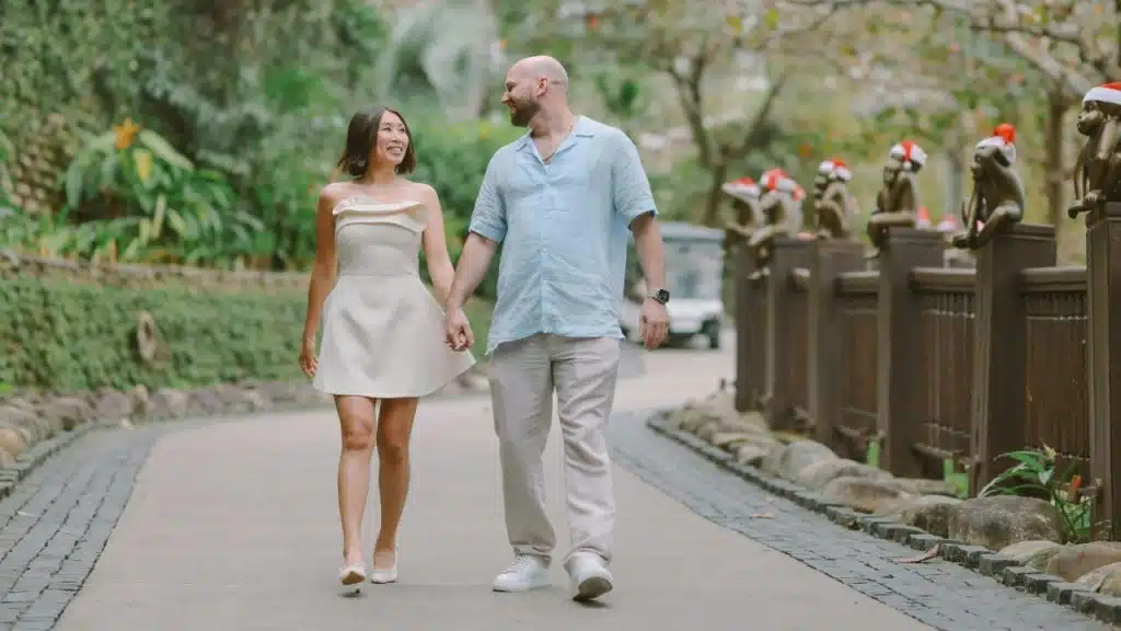 A man and woman hold hands while walking on a path lined with monkey statues.