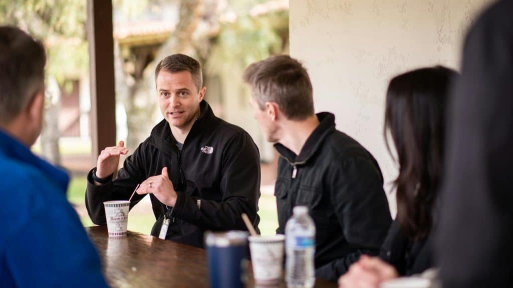 A man speaking to a small group seated around a table outdoors.