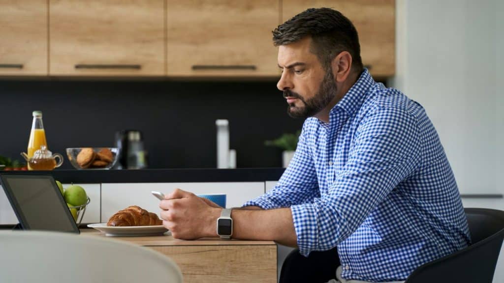 A man sits at a kitchen table looking at a phone and a laptop screen.