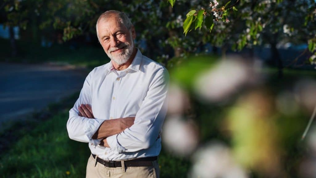 An older man in a white shirt stands outdoors with arms crossed, smiling slightly.