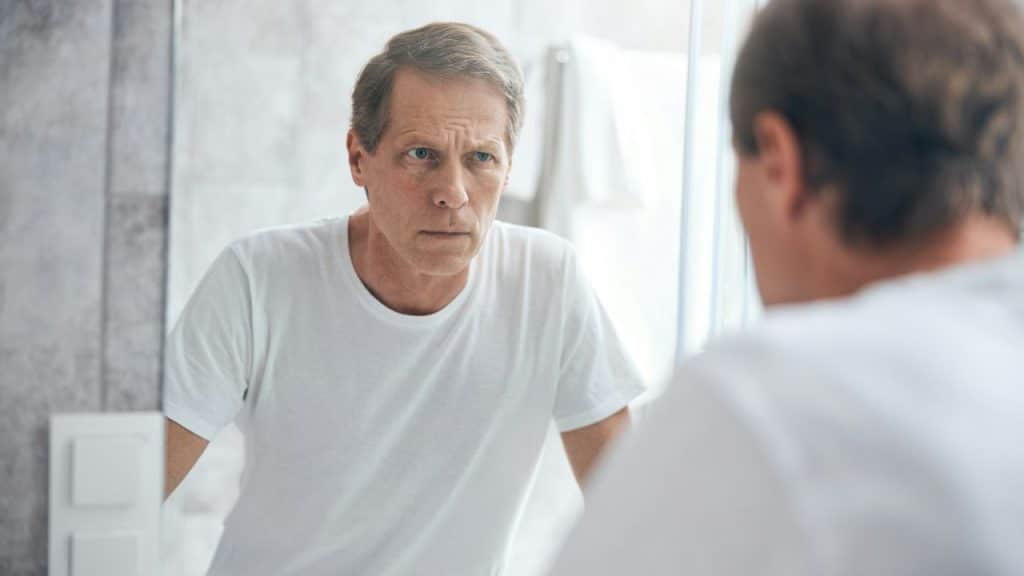 A man in a white t-shirt looks at his own reflection in a bathroom mirror.