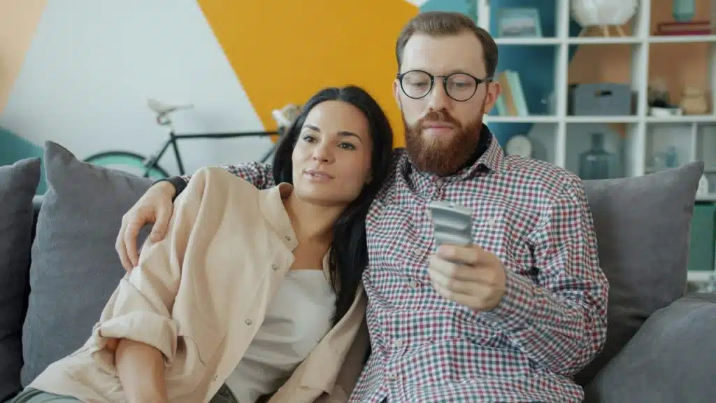 A man with a beard and glasses holds a remote while a woman leans back.