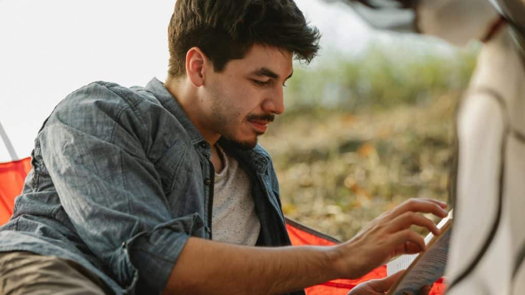 A man with a mustache sits outdoors and looks down at the pages of a book.