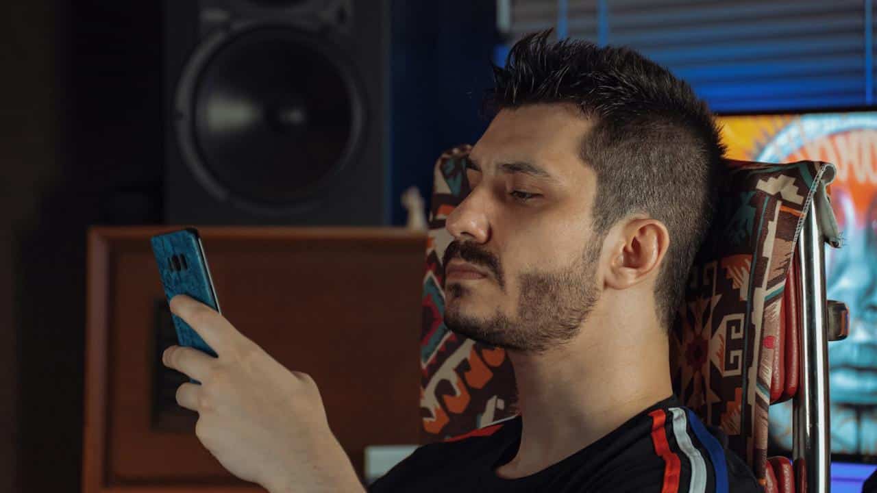 A lightly bearded man looking at his phone with an inscrutable expression. 