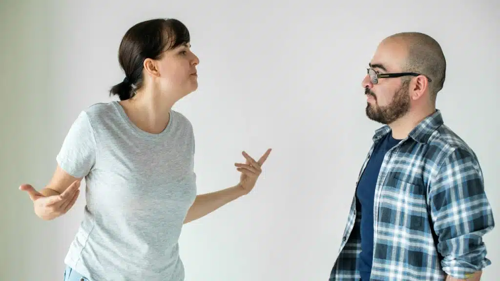 A woman in a grey t-shirt gestures with open hands while facing a man in glasses.