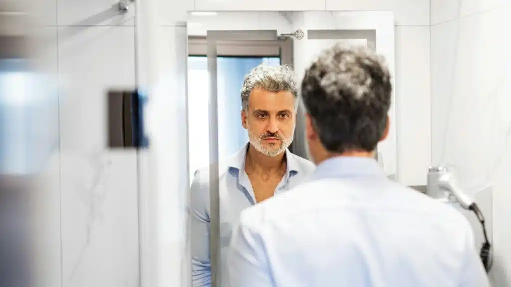 A silver-haired man in a blue shirt is looking intently at his reflection in a bathroom mirror.