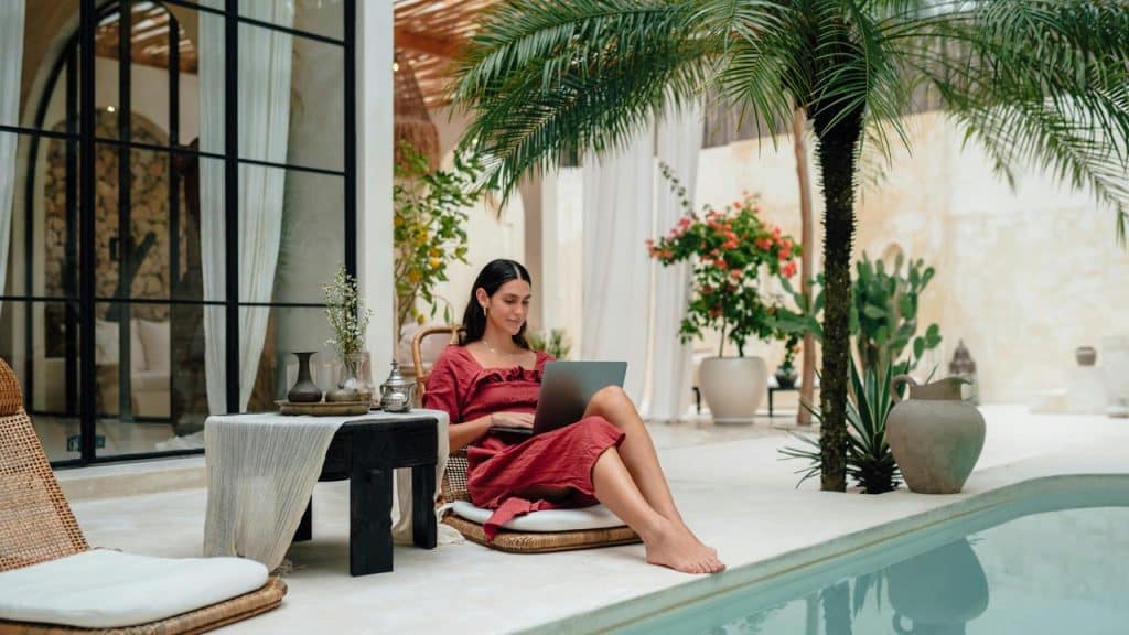 A woman in a red dress working on a laptop beside a pool in a lush courtyard.
