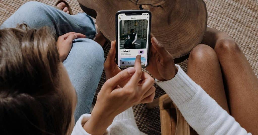 A person holding a phone and liking a dating app profile while sitting with someone nearby.