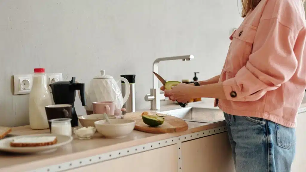 A person in a pink jacket slicing fruit in a kitchen with breakfast items on the counter.