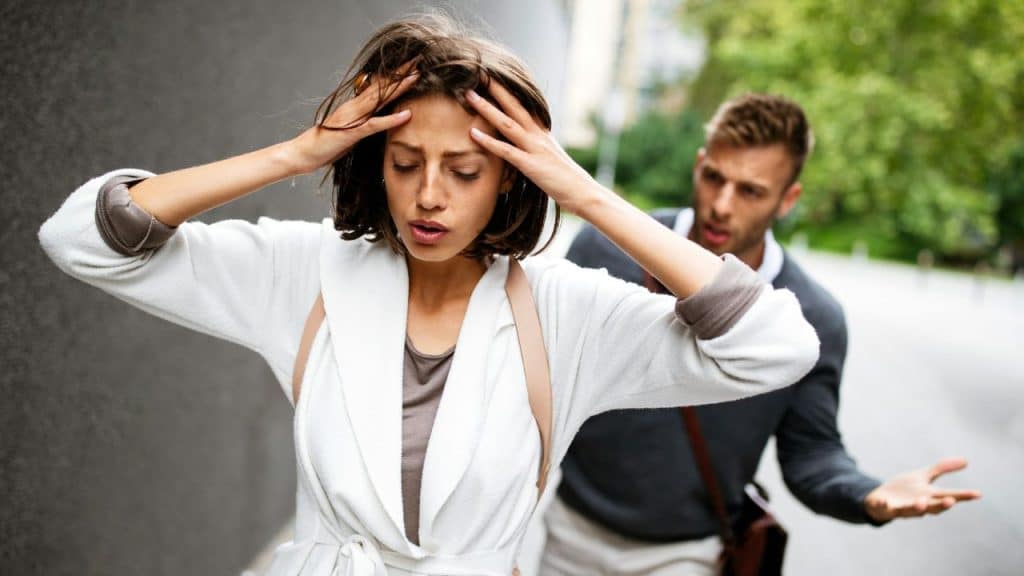 A woman holds her head with both hands while a man gestures toward her in the background.