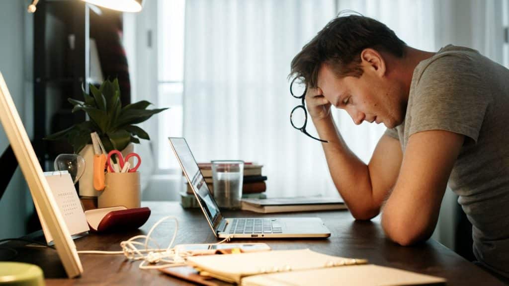 A man leans over a laptop at a desk with his hand on his forehead.