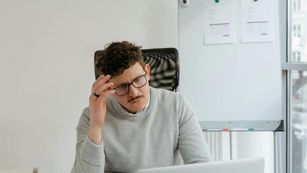 A man sitting at a desk looking at his laptop with his hand on his forehead.