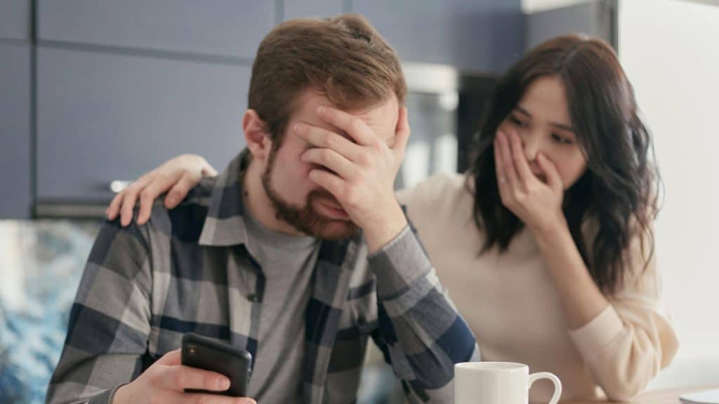A man covers his face while holding a phone as a woman watches with a hand over her mouth.