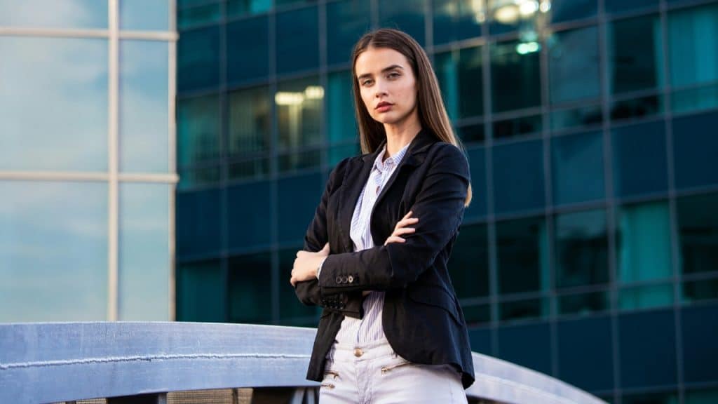 A woman in a black blazer stands with arms crossed in front of a building.