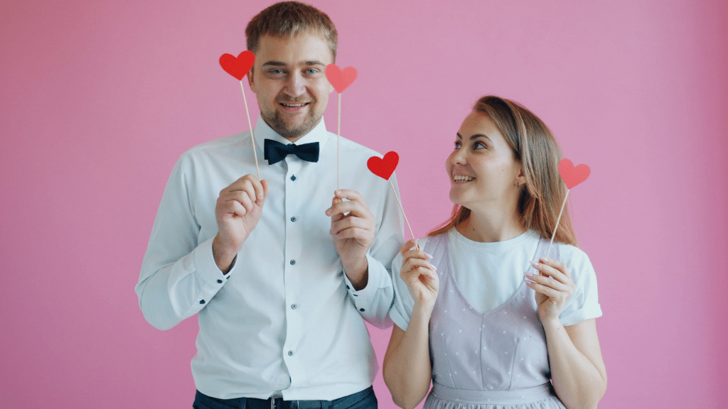 Couple holding red hearts on a stick while they are standing against a pink wall.