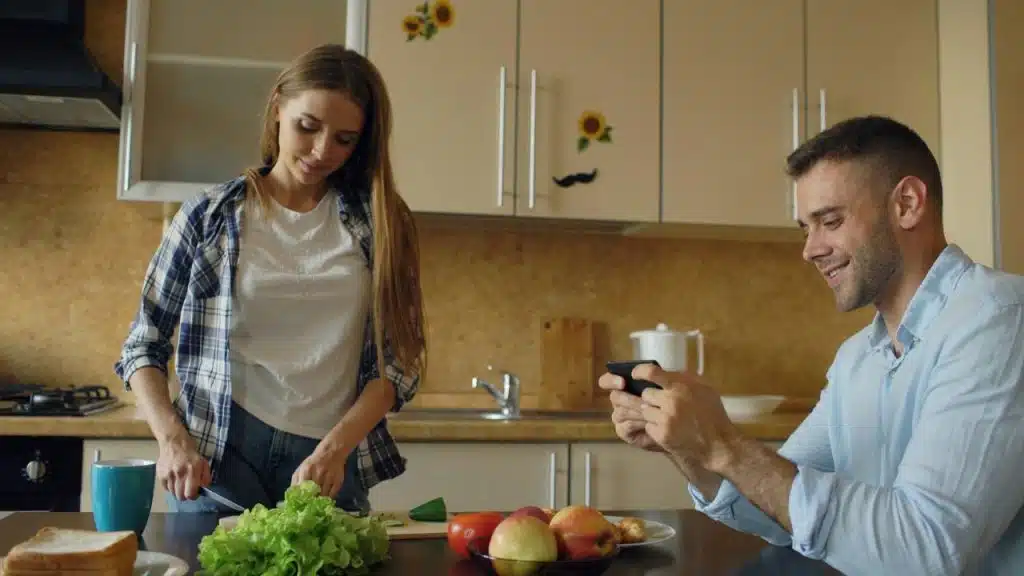 A woman prepares food in a kitchen while a man sitting nearby looks at his phone.