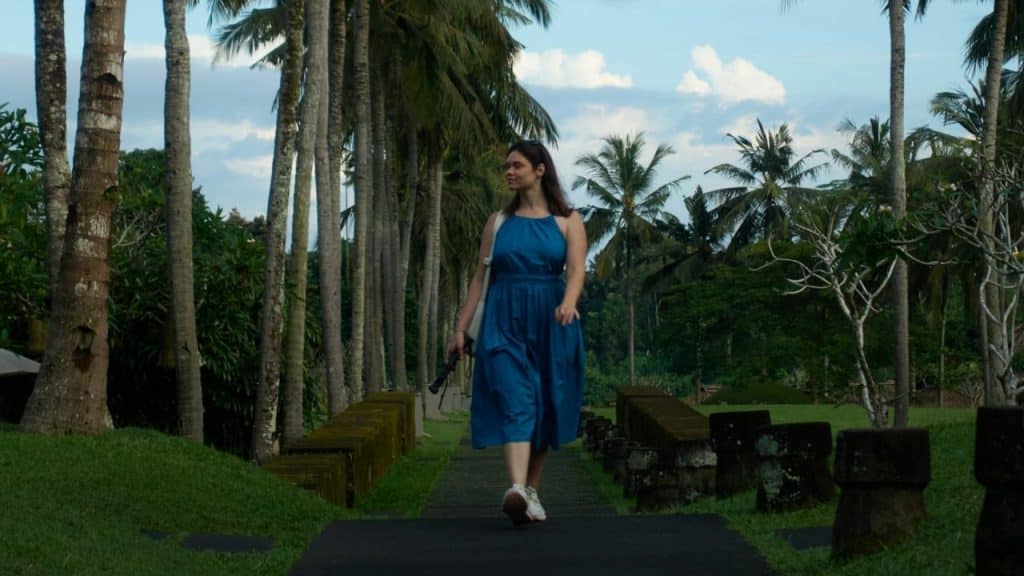 A woman in a blue dress walks down a path lined with palm trees and stone.