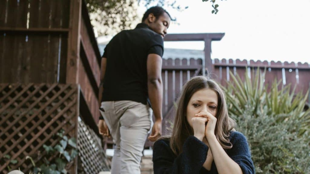 A woman sits in the foreground holding her hands to her mouth while a man walks away.