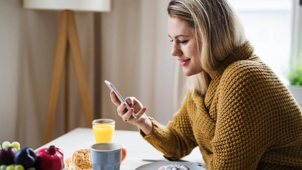 A woman sits at a table, smiling while looking at her smartphone beside breakfast items.