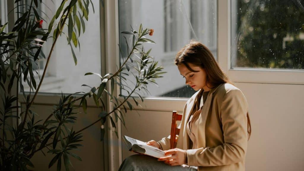 A woman reading a book by a window with sunlight streaming in.