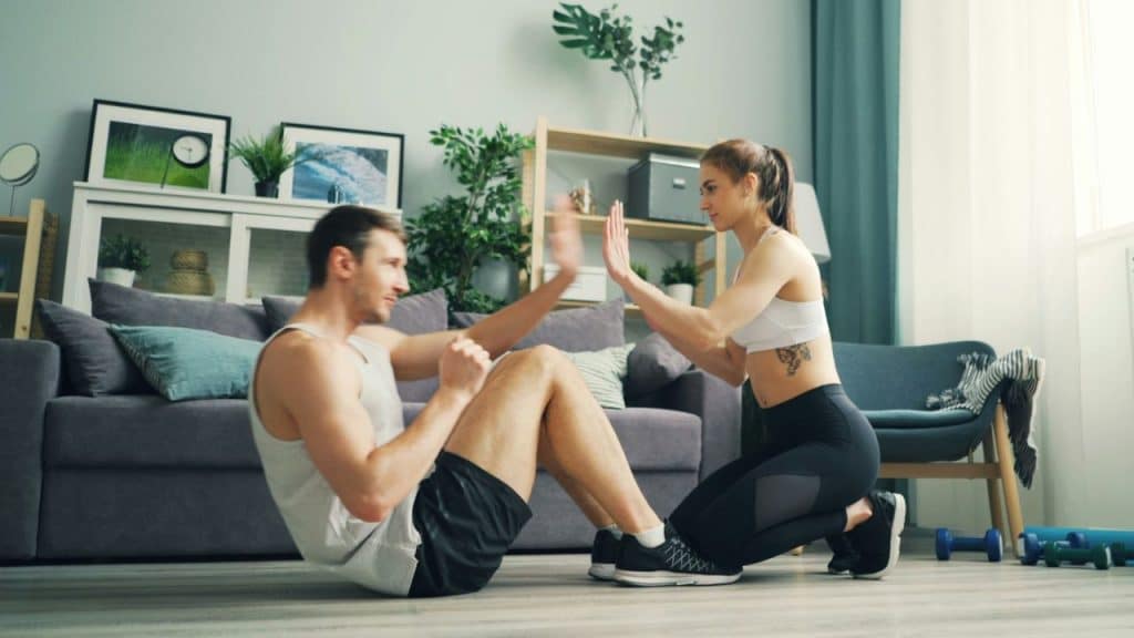 A man performs a sit-up and high-fives a woman kneeling on the floor in a living room.