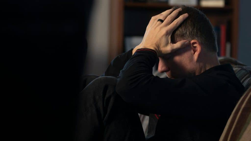 A man in a black shirt sits on a couch with both hands on his head.