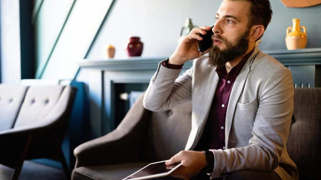 A man sitting indoors, talking on a phone while holding a tablet.