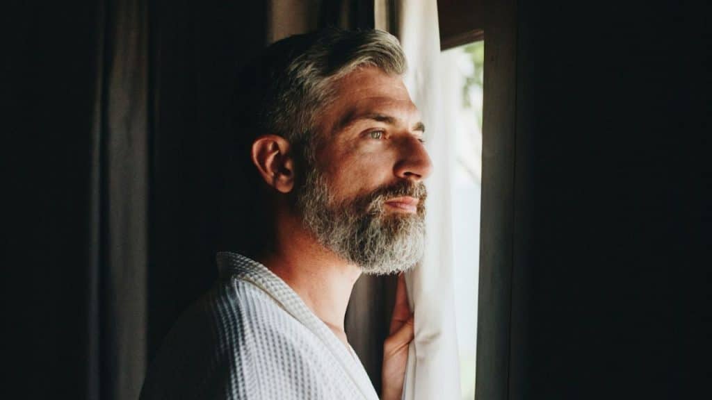A bearded man in a white robe looks out a window through dark grey curtains.