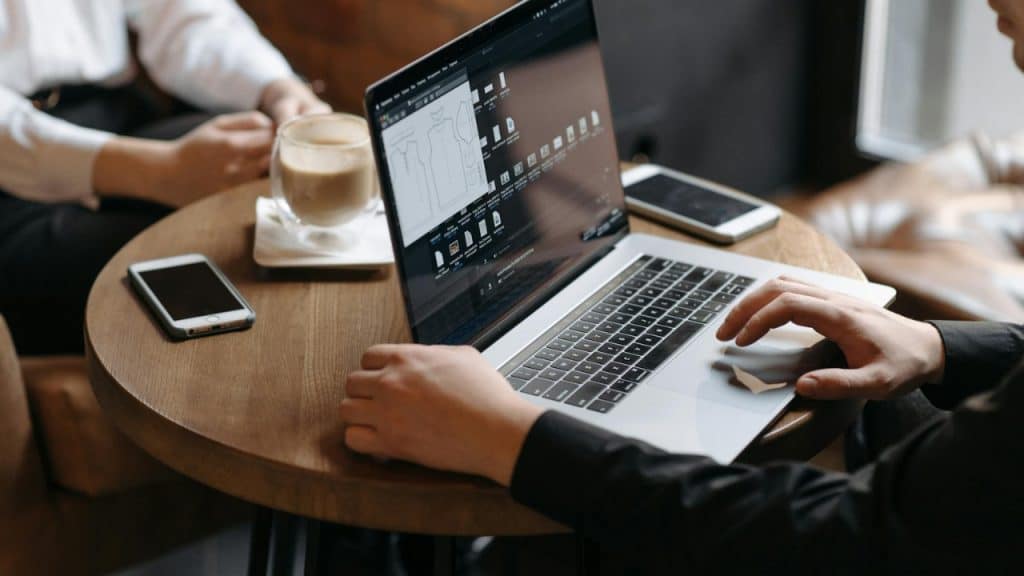 Hands type on a laptop at a cafe table next to smartphones and a coffee.