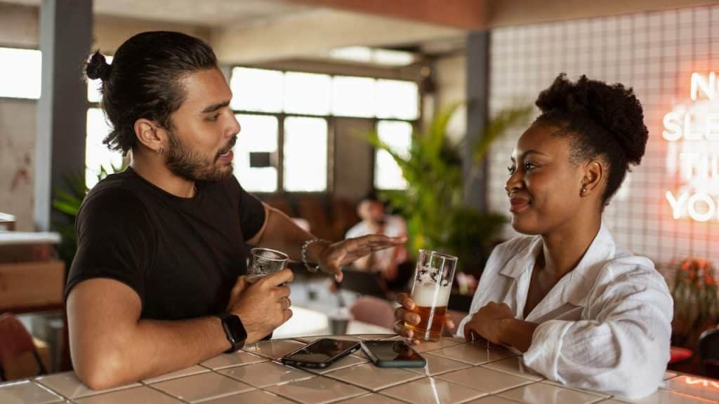 A man with a bun and a woman talk over drinks at a tiled bar.