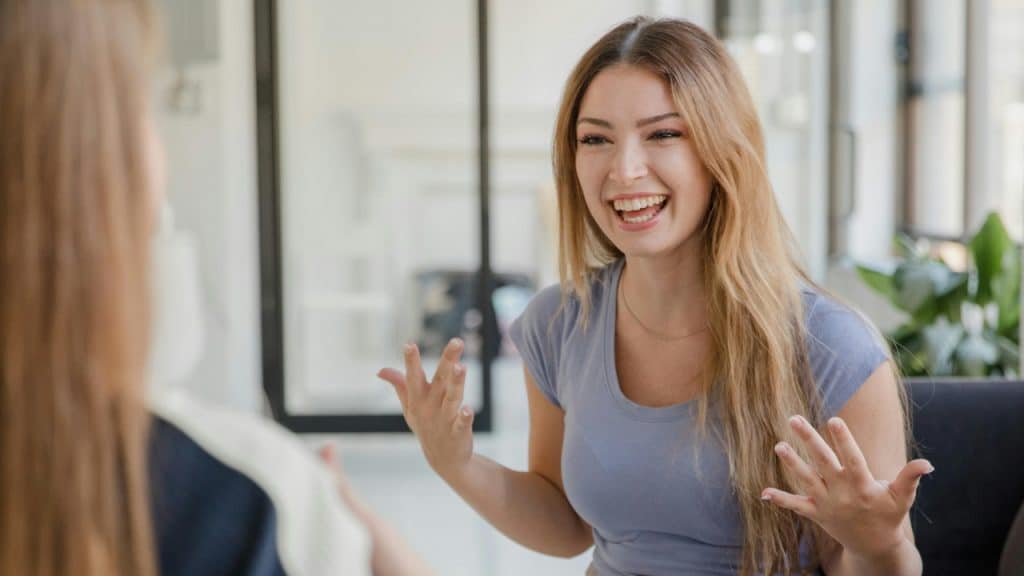 A smiling woman with long blonde hair gestures with her hands while talking to someone.