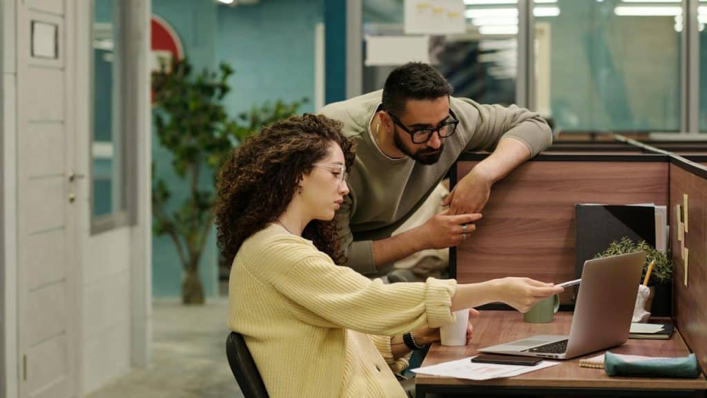 A woman points at a laptop screen while a colleague leans over her desk in an office.