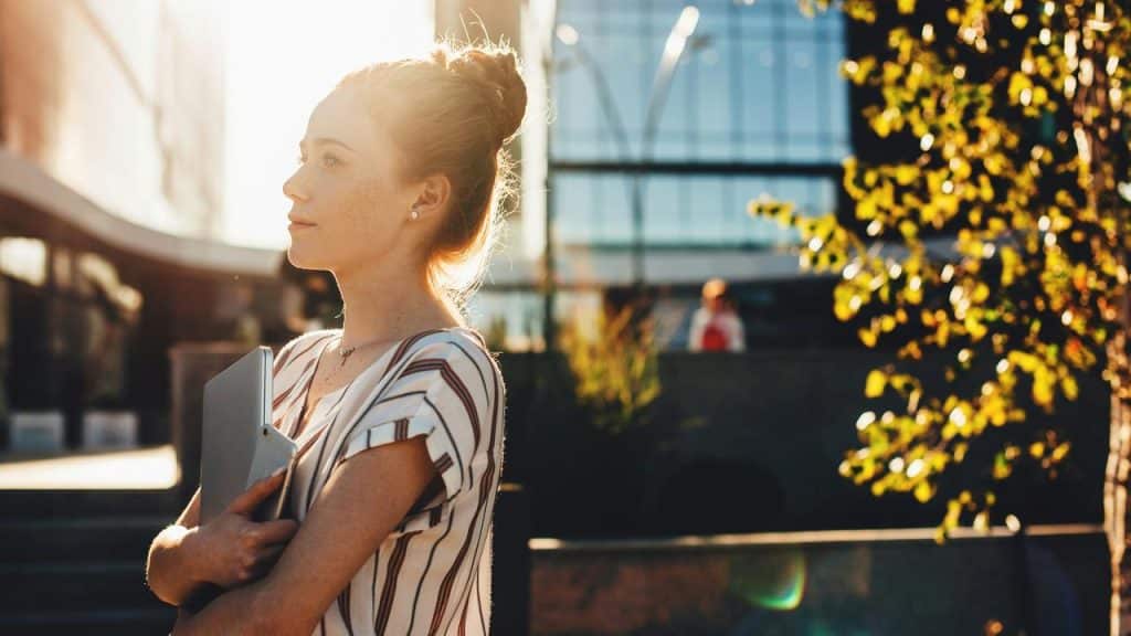 A young woman holding a tablet and looking ahead in warm outdoor light.