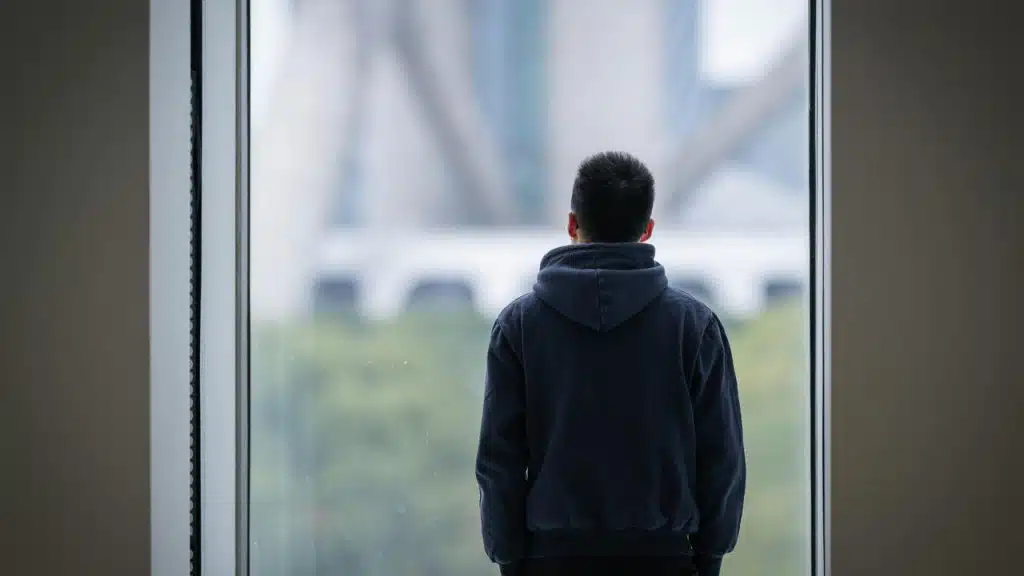 A person in a hoodie stands by a large window looking outside.