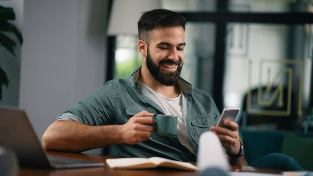 A man smiling while holding a phone and a coffee cup.