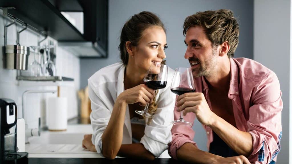 A man and woman in a kitchen smile at each other while holding glasses of wine.