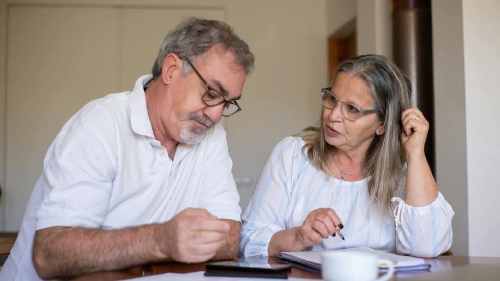 An older man and woman sit at a table looking down at papers and a phone.