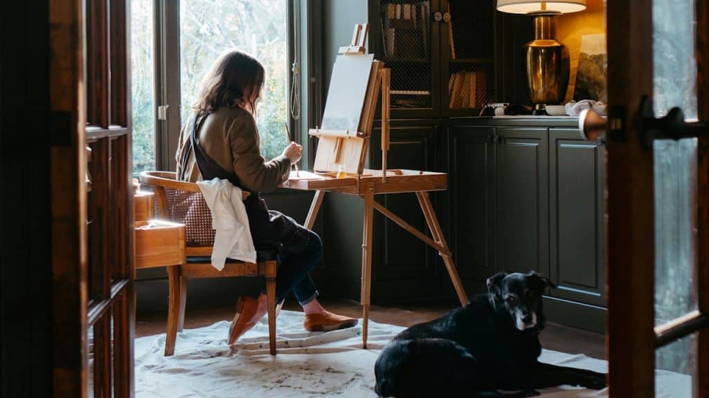 A woman is painting while her dog sits on the floor near her.