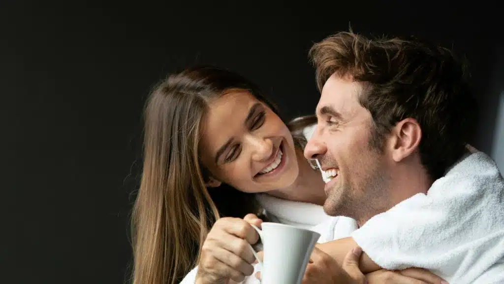 A smiling couple in bathrobes cuddling while holding a cup.
