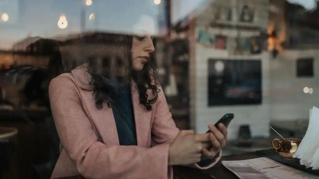 A woman in a pink coat sits by a window, looking at her phone in a café.