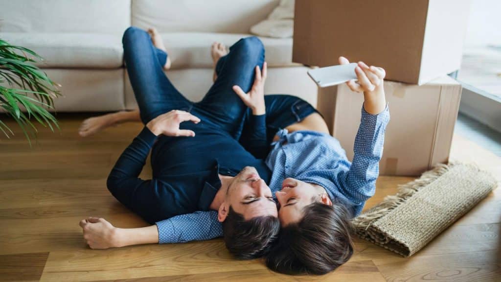 A couple lies on the floor taking a selfie near moving boxes.